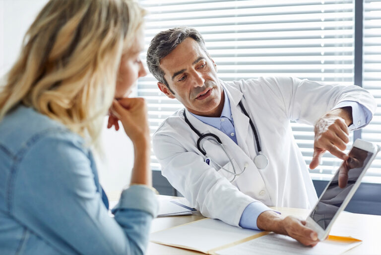 Ein Arzt sitzt am Schreibtisch gegenüber einer Patientin. In der rechten Hand hält er ein Tablet und zeigt mit dem Finger auf das Display. Er erklärt der Frau offenbar wichtige Sachverhalte.