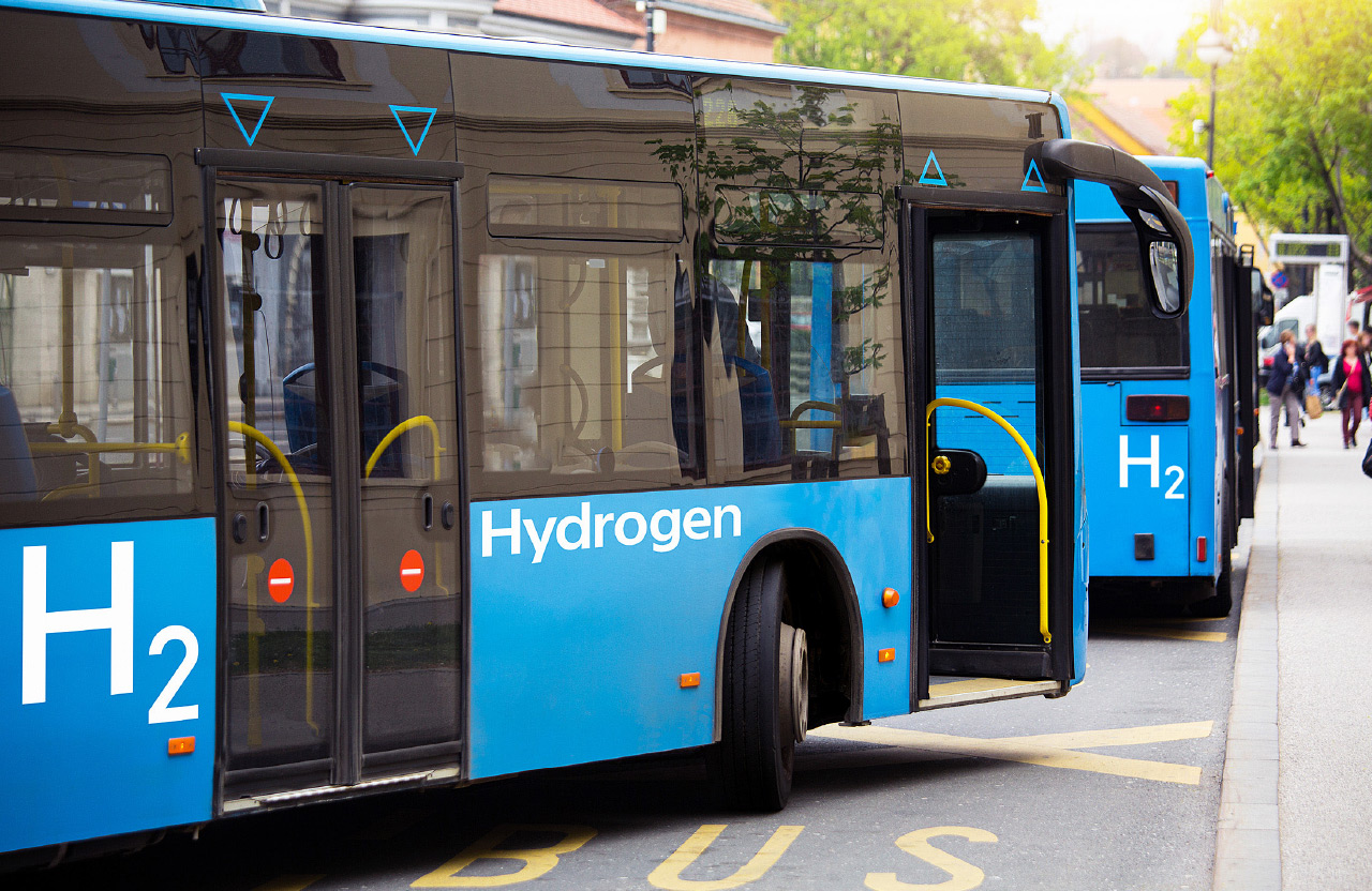 Zwei Blaue Wasserstoff-Linienbusse im Stadtverkehr, die weiß mit H2 und Hydrogen beschriftet sind.