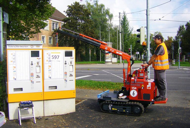 Vor einer Kreuzung stehen zwei etwa mannshohe Fahrscheinautomaten: Einer der Automaten hängt bereits am Ausleger eines kleinen roten Raupenfahrzeug, dass von einem Arbeiter gesteuert wird. Vorne auf dem Fahrzeug ist eine kleine Ladefläche erkennbar, auf der der Automat abgstellt werden kann.