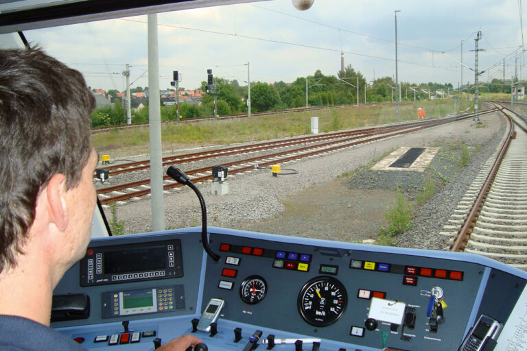 Blick aus dem Führerstand eine Lokomotive: Zu sehen sind innen Tacho, diverse rote, gelbe, blaue und weiße Leuchtschalter, ein Handy im Halter, ein Mikrofon und Schalttafeln mit kleinen Displays. Aus dem Fenster sind neben dem eigenen Fahrbett noch zwei doppelgleisige Strecken zu sehen, Oberleitungen und Signale.