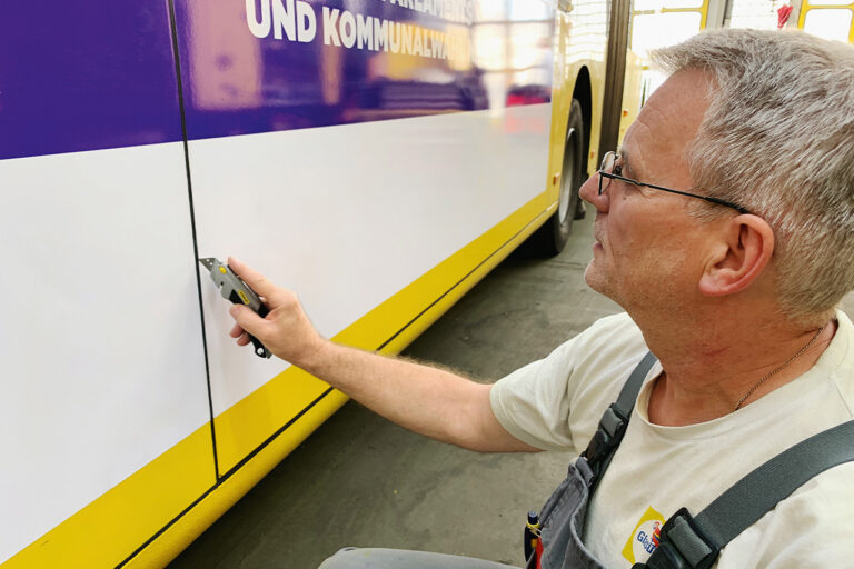 Ein Manns schneidet mit einem Cuttermesser konzentriert die Folie auf der Seite eines Omnibusse an der Nahtstelle zur mittleren Tür zu.