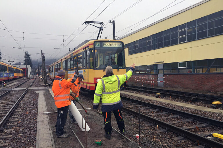 Drei Mitarbeiter im Gleisbereich in Signalkleidung geben einer heranfahrenden Tram Handzeichen.