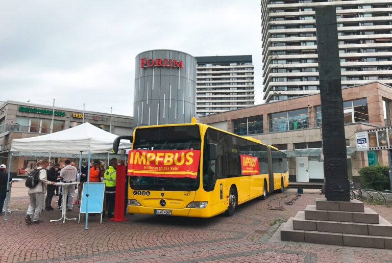 Gelber Impfbus von schräg vorn auf einem Platz vor einer Einkaufspassage, mehrere Menschen unter einem Zeltdach beim Anmelden zur Impfung.