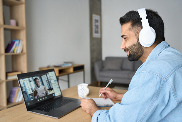 Mann mit Kopfhörern sitz am Tisch vor einem aufgeklapptem Laptop und hat einen Stift in der Hand und einen Schreibblock vor sich.