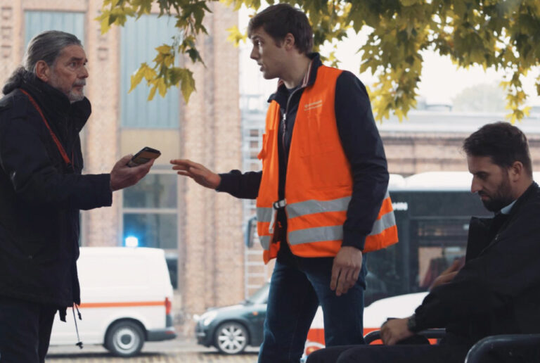ein Mann sitzt trübsinnig auf einer Bank, davor steht ein Mann in einer orangenen Warnweste und hält einen dritten Mann von dem sitzenden Mann fern