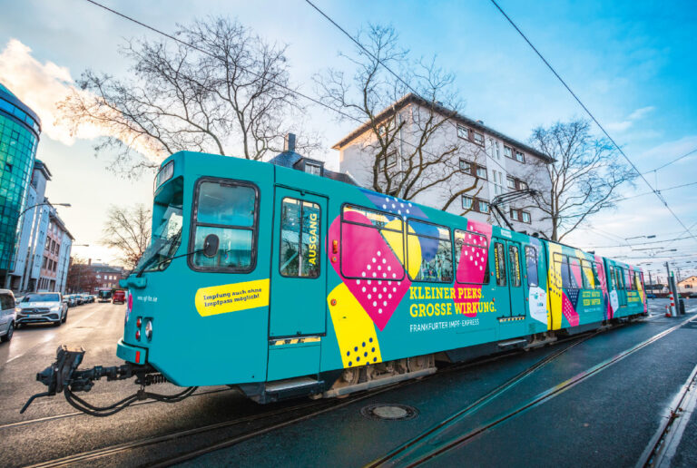 Türkisfarbene Straßenbahn von der Seite, mit bunter Aufschrift „Kleiner Pieks, große Wirkung“ im Stadtgebiet