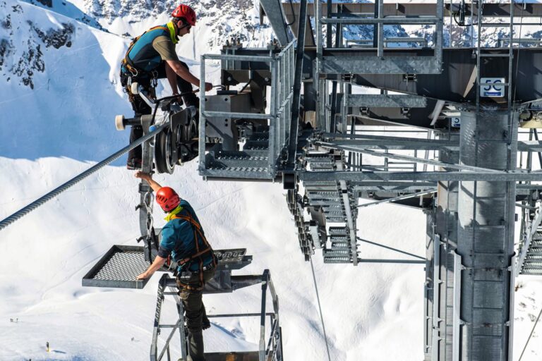 Zwei Mitarbeiter mit Gurten und Helmen gesichert bei der Wartung einer Seilbahn in großer Höhe vor winterlicher Bergkulisse